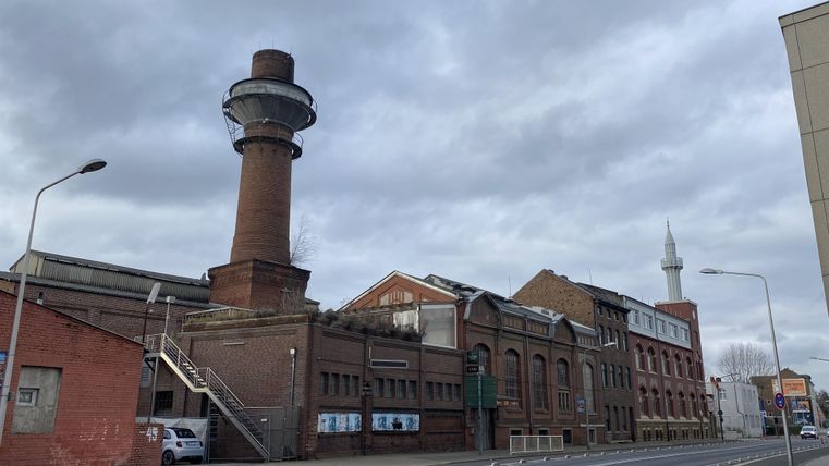Ein urbaner Straßenzug mit historischen Gebäuden und einem markanten Aussichtsturm. Der Himmel ist bewölkt und die Straßen sind ruhig.
