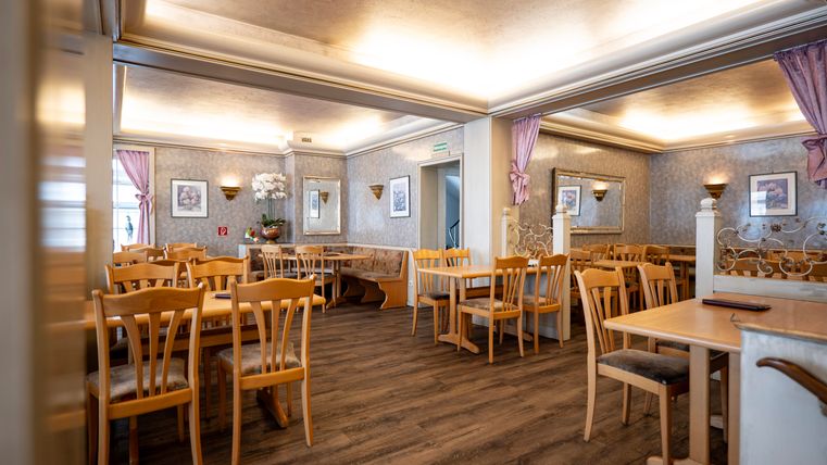 A cozy restaurant with wooden chairs and tables. The walls are adorned with decorative pictures and curtains.