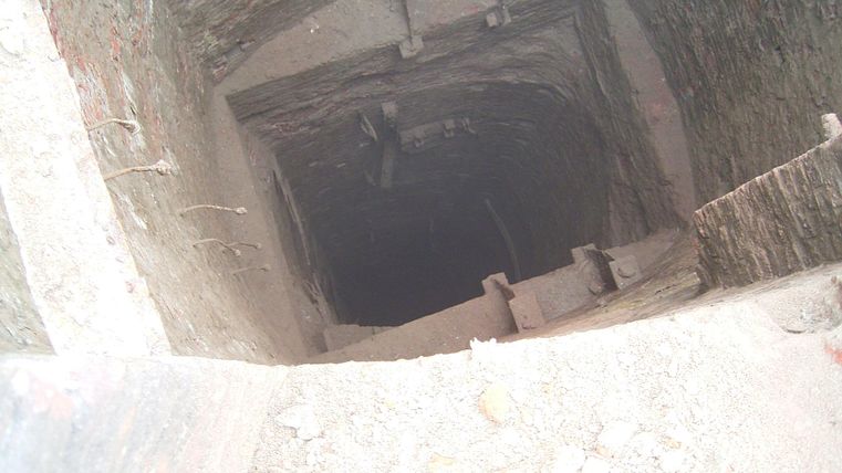 Ein dunkler Schacht mit steilen Wänden und wenigen sichtbaren Treppenstufen. Der Boden ist mit losem Material bedeckt.