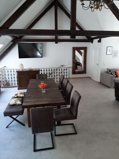 Een moderne eethoek onder een hoge dak met zichtbare houten balken. De tafel is gedekt en er zijn comfortabele stoelen evenals een gezellige bank in de hoek.