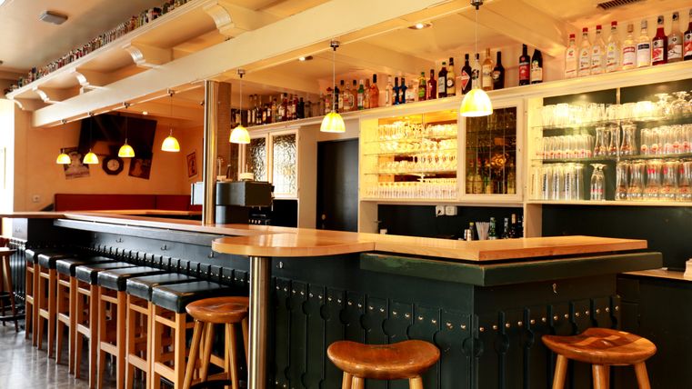 A modern bar with wooden chairs and a long counter. The shelves are filled with various drinks and glasses.