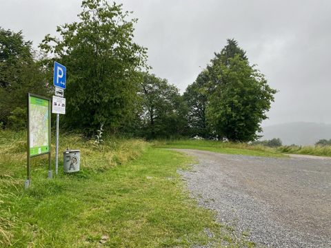 A parking lot in nature, surrounded by trees and grass. The sky is cloudy, and the path leads into the greenery.
