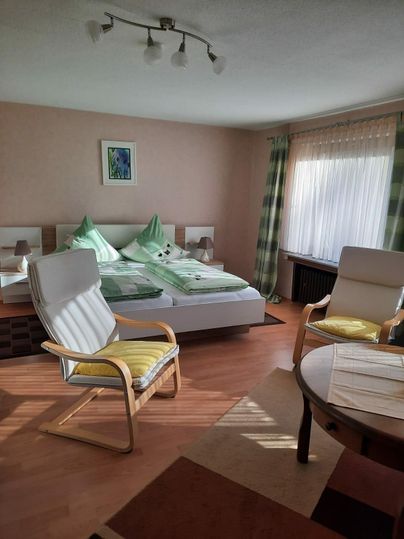 Een gezellige slaapkamer met een groot bed en twee stoelen. De muren zijn in zachte kleuren geschilderd en veel daglicht valt door de ramen.