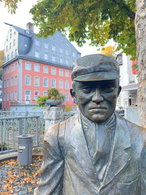 Een bronzen beeld van een man in een pak en een hoed staat voor een historisch gebouw. Op de achtergrond zijn meer oude gebouwen en bomen te zien.