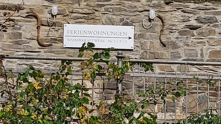 Een bord met de tekst "Vakantiewoningen" aan een stenen muur. Er zijn planten en een richtingwijzer die naar een accommodatie wijst.