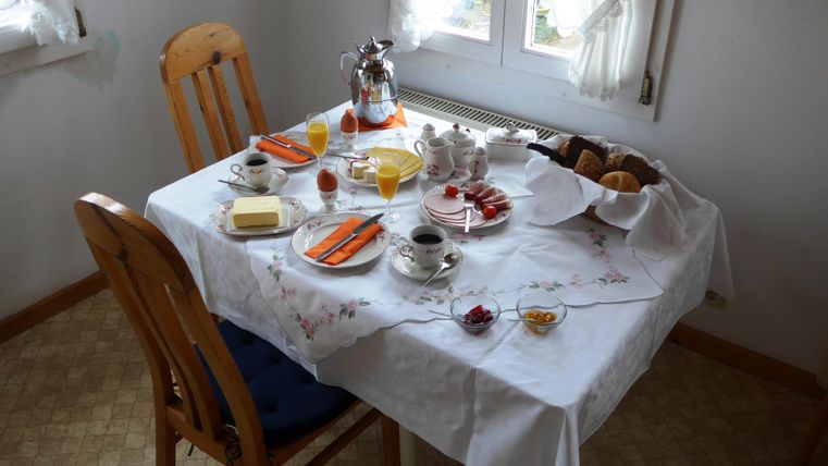 A lovingly set breakfast table with various treats. There are rolls, jam, fruit, and drinks on it.