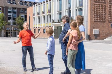 Eine Gruppe von fünf Personen steht in einem öffentlichen Raum. Sie scheinen miteinander zu sprechen und zu lachen, während im Hintergrund moderne Gebäude sichtbar sind.