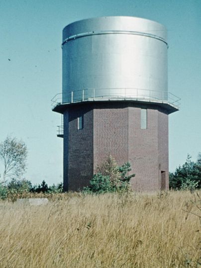 Ein hoher Wasserturm aus Ziegelsteinen mit einem metallenen oberen Teil. Er steht in einer offenen, grünen Landschaft mit hohem Gras.