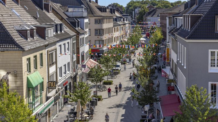 Eine belebte Einkaufsstraße mit Bäumen und Geschäften. Viele Menschen schlendern entlang der Straße.