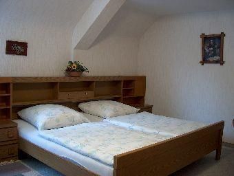 A simple bedroom with a double bed and two pillows. Pictures hang on the wall, and a small plant is on the nightstand.