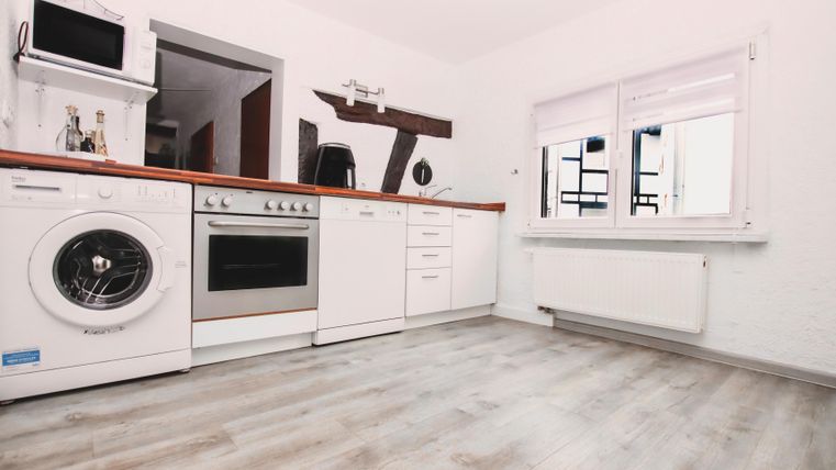 Een moderne keuken met een wasmachine, oven en witte kast. De vloer is van licht hout en er is een groot raam.