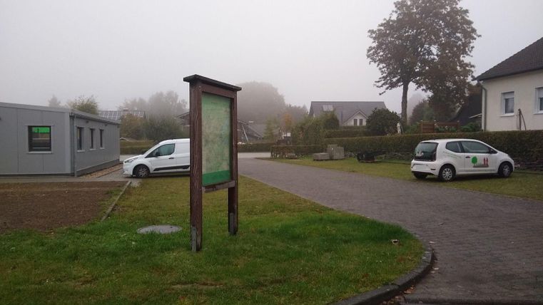 A foggy place with an information board and two cars in the parking lot. In the background, some buildings and trees are visible.