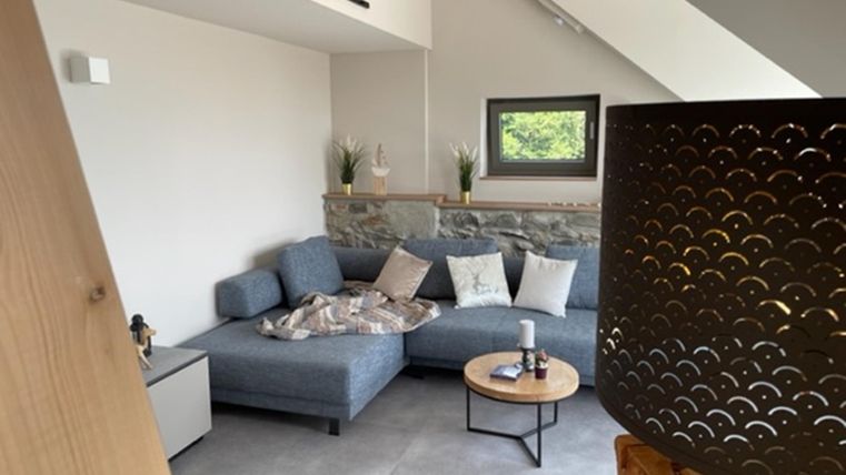 Een moderne woonkamer met een grijze bank en een ronde tafel. De inrichting is helder en vriendelijk vormgegeven, met planten en houten accenten.
