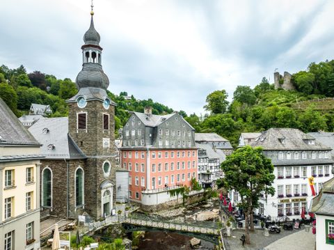 Een pittoresk stadsplein met een kerk en historische gebouwen. Op de achtergrond is een heuvel met ruïnes te zien.