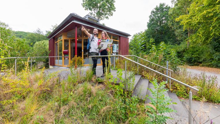Ein Besucherzentrum mit einer roten Fassade inmitten von Bäumen und Pflanzen. Zwei Personen schauen auf eine Karte und genießen die Umgebung.
