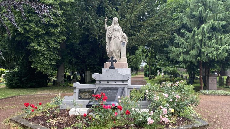 Eine Statue steht inmitten eines freundlich gestalteten Grabes, umgeben von bunten Blumen. Im Hintergrund sind hohe Bäume sichtbar, die eine ruhige Atmosphäre schaffen.