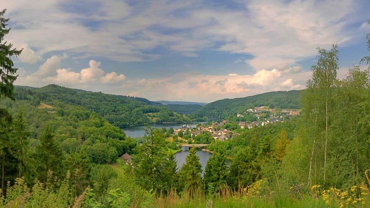 Eine malerische Landschaft mit sanften Hügeln und einem Fluss. Der Himmel ist teils bewölkt und die Umgebung ist üppig grün.