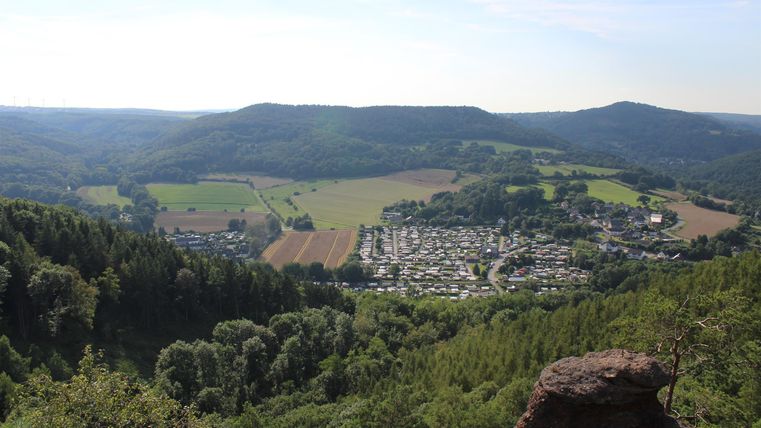 Eine malerische Aussicht auf ein Tal mit einem kleinen Dorf und umgeben von grünen Hügeln. Der Himmel ist klar und die Landschaft strahlt Ruhe aus.