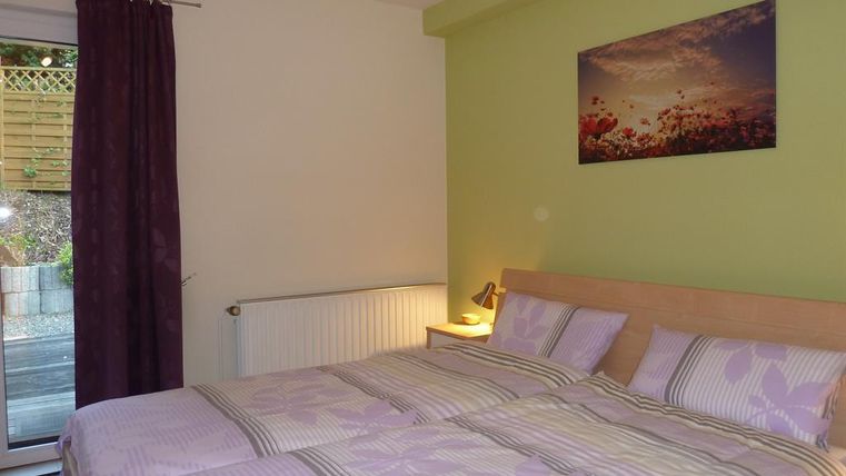 A cozy bedroom with two beds and purple bedding. A picture of a flower landscape hangs on the wall, and there is a window with a view to the outside.