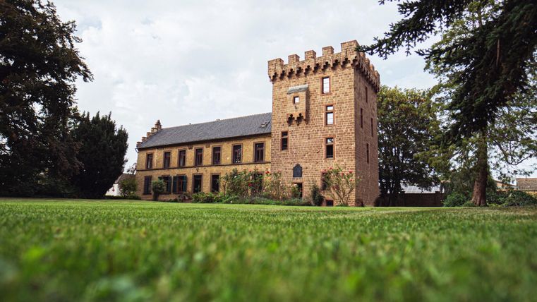 Een historisch gebouw met een vierkante toren en grote vensters. De omliggende tuin is groen en goed onderhouden.
