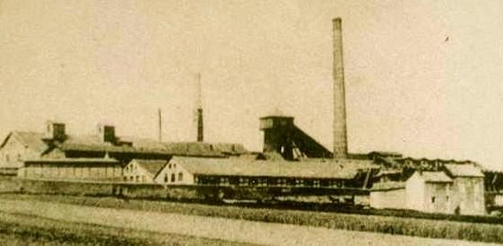 Een oude fabriek met hoge schoorstenen en verschillende gebouwen. De omgeving is landelijk en uitgestrekt.