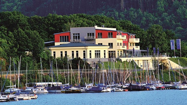 Ein modernes Gebäude am Wasser mit vielen Segelbooten im Vordergrund. Die Umgebung ist grün und bietet eine schöne Aussicht auf die Natur.