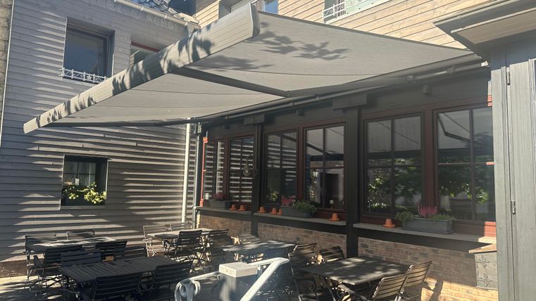 A terrace with a large awning and several tables. The restaurant is surrounded by a beautiful building.