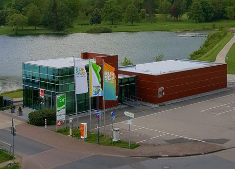 A modern building by the water with large windows. In the foreground, flags and a paved area can be seen.