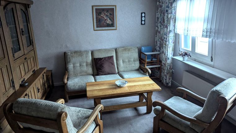 A cozy living room with a wooden bench, a table, and simple furniture. The light gently filters through a window, and there is a wall decoration.