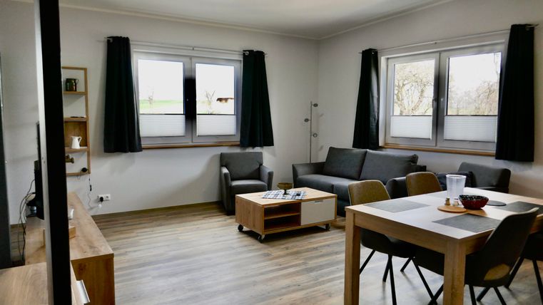 A modern living room with a gray sofa, a coffee table, and a dining table. Large windows let in plenty of light, and the walls are painted in neutral colors.
