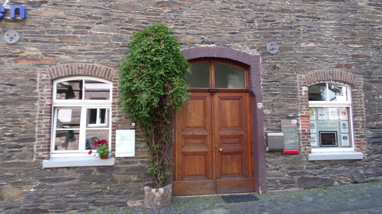 Een oude stenen muur met een grote houten deur in het midden. Links en rechts zijn er ramen met bloemen op de vensterbanken.