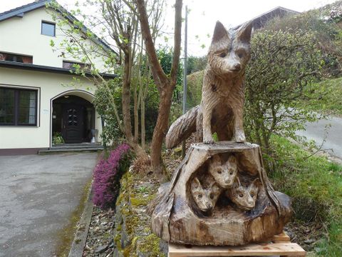 A wooden sculpture of a fox and its cubs stands in front of a house. The statue is detailed and portrays the animals in a natural environment.