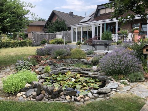 Ein schöner Garten mit einem Teich, umgeben von bunten Pflanzen und Lavendel. Im Hintergrund steht ein Haus mit einer überdachten Terrasse.