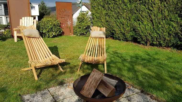 Een mooie tuin met twee houten ligstoelen en een vuurplaats. De omgeving is groen en uitnodigend.