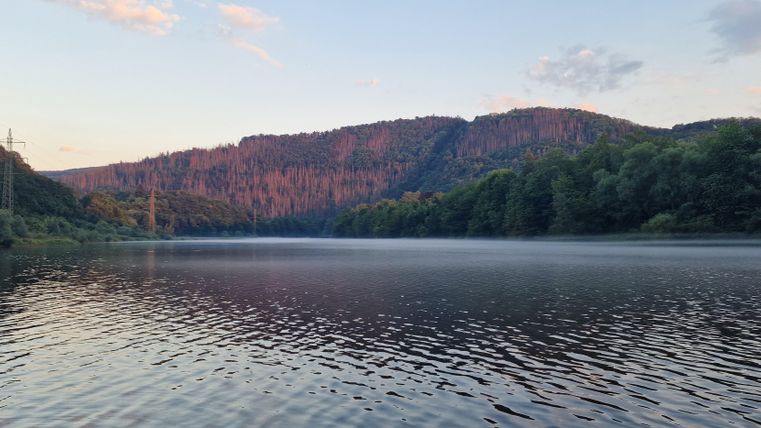 A tranquil lake surrounded by wooded hills. The sky is lightly clouded and the light of dusk reflects in the water.