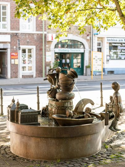 An artistic fountain with figures drawing water from various basins. Surrounded by trees and near a building in the city.