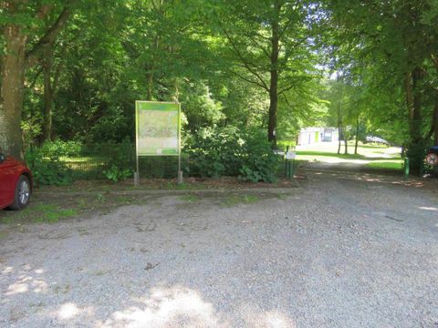 Ein schmaler Schotterweg zwischen Bäumen führt zu einem Parkplatz. Im Hintergrund sind weitere Bäume und ein Schild sichtbar.