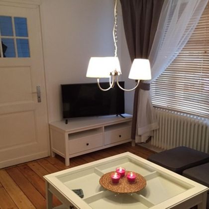 A cozy living space with a television, a coffee table, and a modern lamp. The windows are equipped with curtains and blinds, and the floor is made of wood.