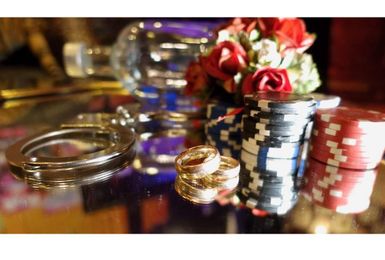 Eine Nahaufnahme von Spielchips in verschiedenen Farben, einem Paar Ringen und einer Handfesseln. Im Hintergrund sind dekorative Blumen zu sehen.