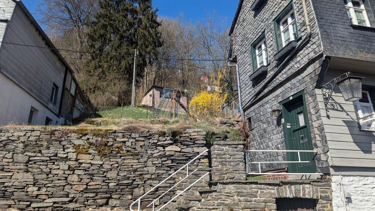 A steep path with a stone wall and two houses. In the background, trees and a meadow with yellow flowers can be seen.