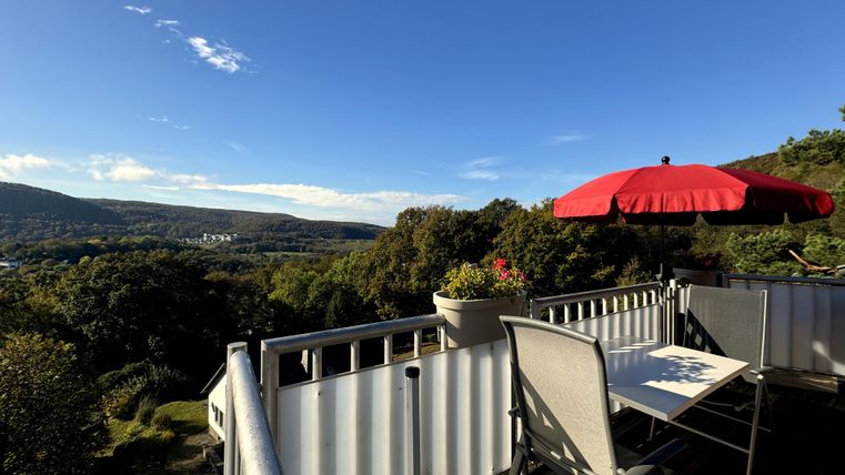 Een balkon met een tafel en stoelen biedt een mooi uitzicht op de omringende bergen en het dal. Een rode parasol staat klaar voor schaduw.
