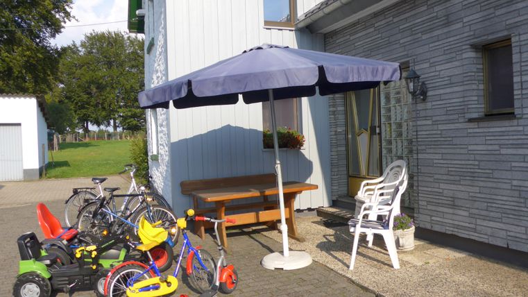 An entrance with a sun umbrella and a table. Next to the entrance, there are various toys and bicycles.