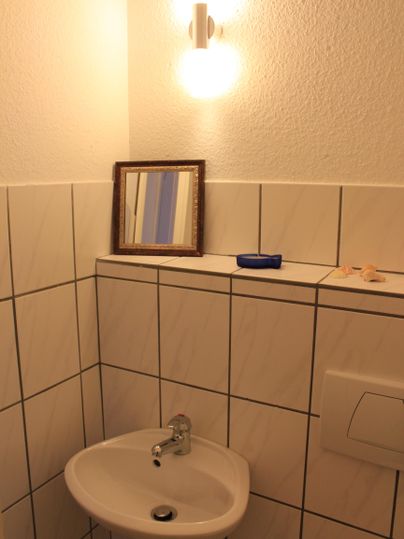 Een kleine badkamer met een wastafel en een wandlamp. Op de plank staan een spiegel en een aantal kleine voorwerpen.