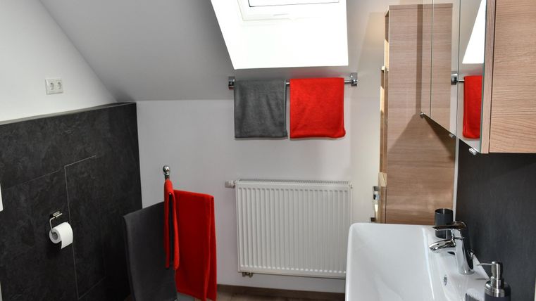 Een modern badkamer met een raam en veel daglicht. De muren zijn in neutrale kleuren gehouden, aangevuld met rode en grijze handdoeken.