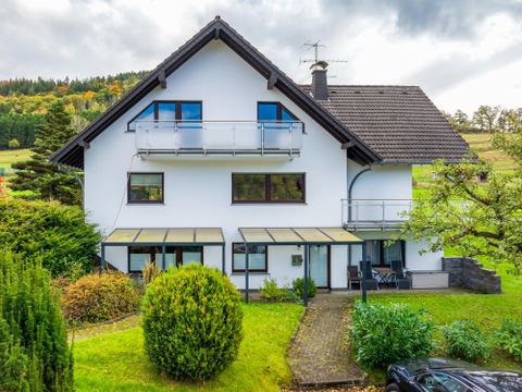Een modern huis met een grote tuin. Het heeft balkons en is omgeven door bomen.