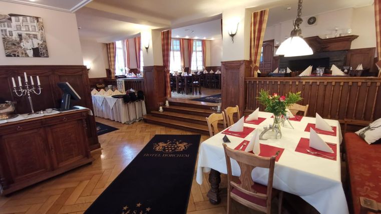 An elegant restaurant with wooden interior and laid tables. In the background, more tables and windows with curtains are visible.