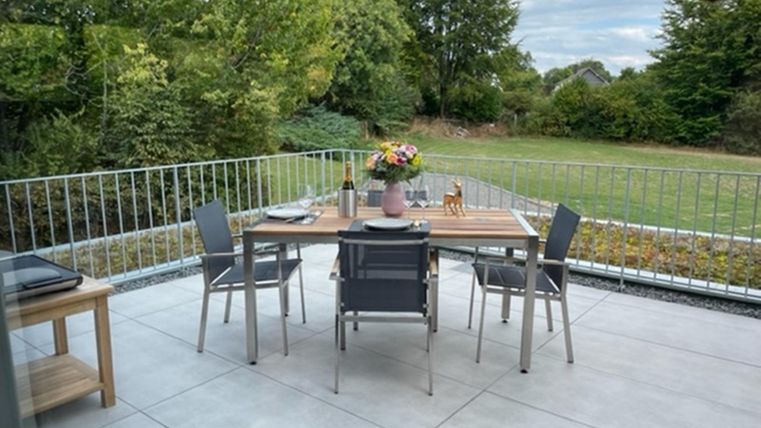 Een elegante eetruimte op een terras met een tafel en stoelen. Uitzicht op een verzorgde tuin met bomen op de achtergrond.