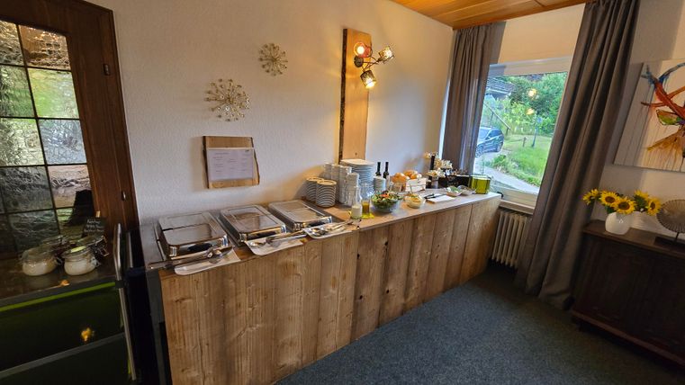 A breakfast buffet with various dishes is set on a wooden table. A green garden landscape can be seen through a window.