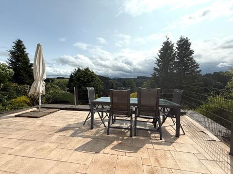 Een terras met een tafel en stoelen, omgeven door groene bomen. De lucht is gedeeltelijk bewolkt en het landschap is goed zichtbaar.