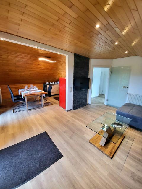A modern living room with a wooden ceiling and a cozy sofa. The dining area is equipped with a table and black chairs.